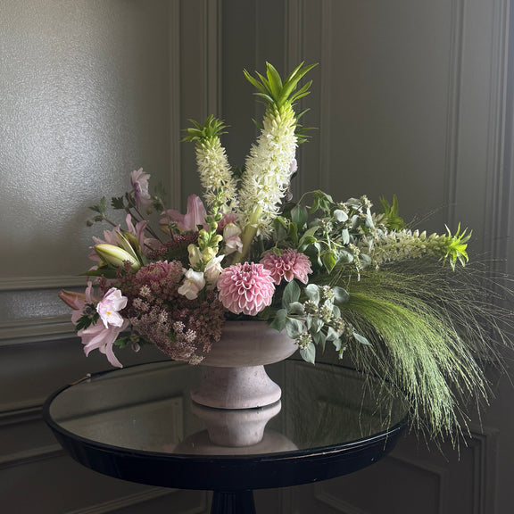A floral arrangement in a pale pink urn-shaped vase, displayed on a round black table against a gray paneled wall.