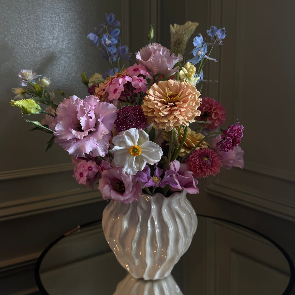 A colorful artificial flower arrangement in a ribbed white ceramic vase, displayed on a dark round table against a dark paneled wall.