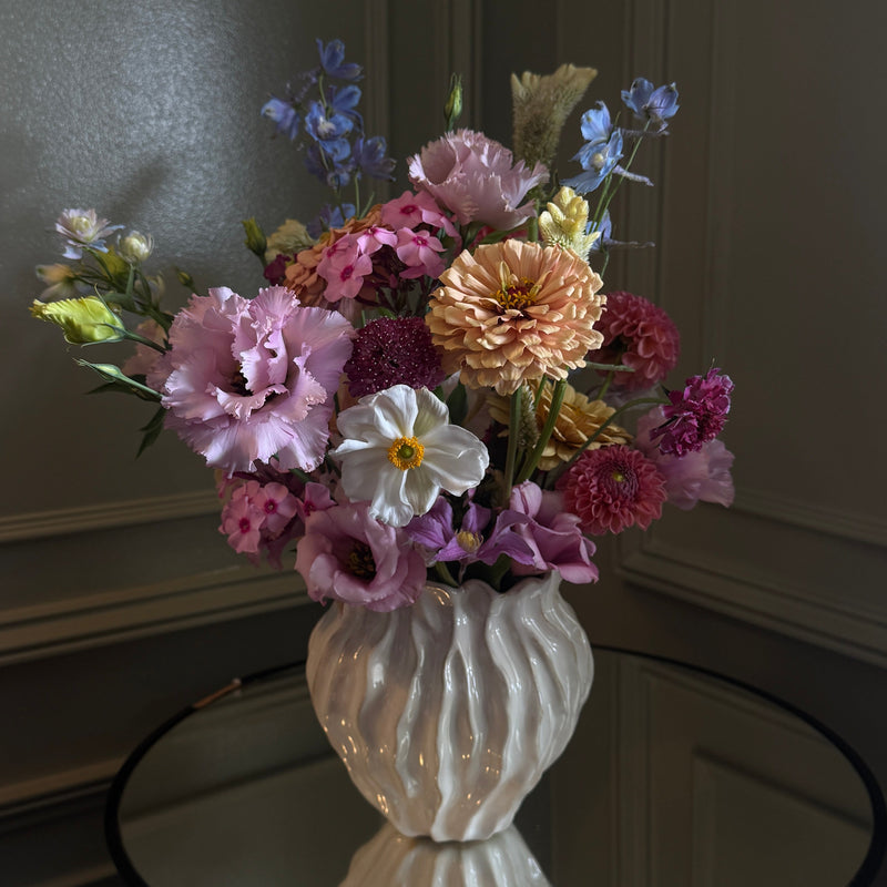 A colorful artificial flower arrangement in a ribbed white ceramic vase, displayed on a dark round table against a dark paneled wall.