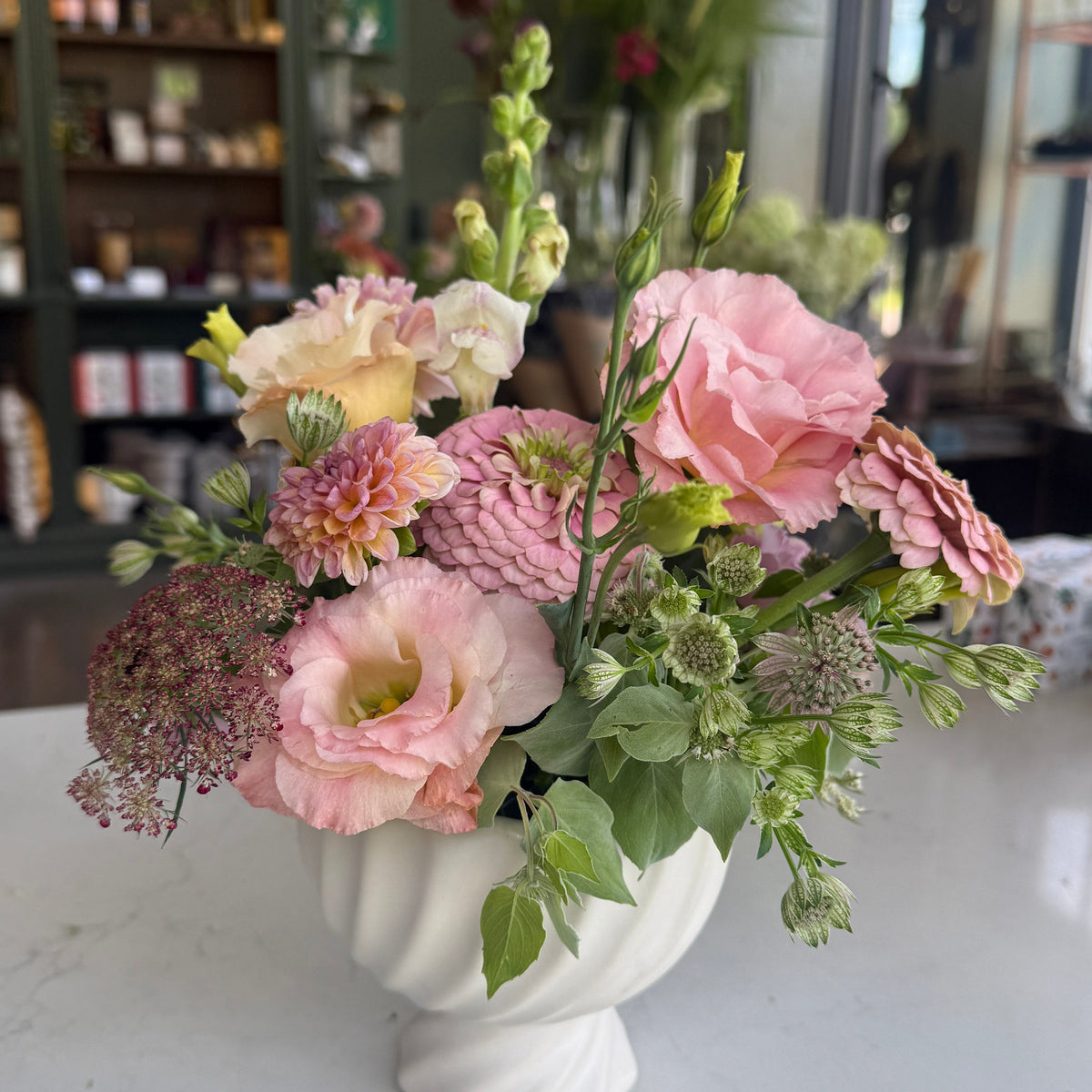 A floral arrangement of pink, peach, and cream flowers with green foliage in a white pedestal vase, set on a light marble countertop inside a softly blurred shop interior.