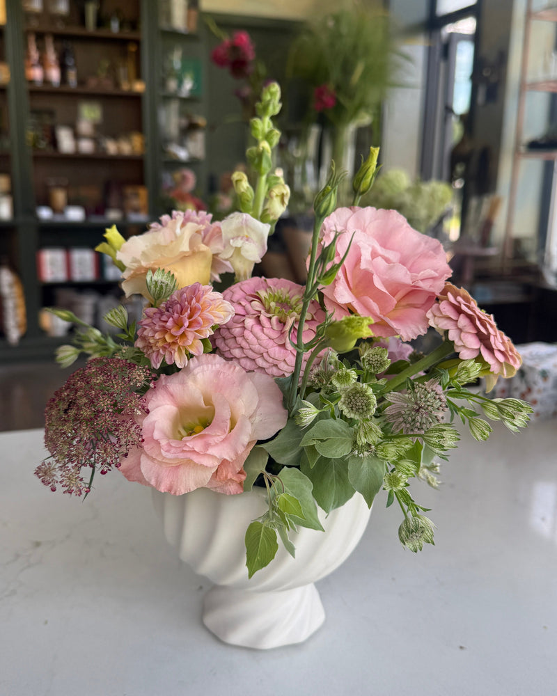A floral arrangement of pink, peach, and cream flowers with green foliage in a white pedestal vase, set on a light marble countertop inside a softly blurred shop interior.