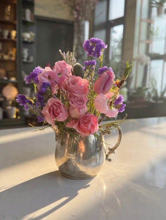 Silver vase with pink and purple flowers on a reflective surface. Flowers are artfully arranged.
